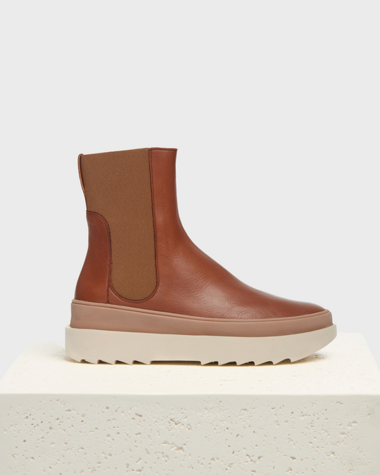 A brown leather ankle boot with a smooth texture, featuring a contrasting elastic side panel, a pull tab at the back, and a thick, beige rubber sole with a ridged pattern.