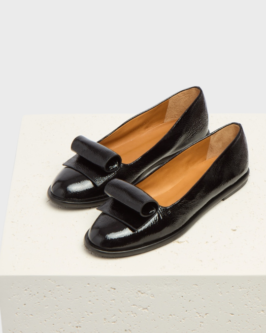 A pair of sleek, black patent leather loafers with a glossy finish, featuring a distinctive folded strap across the top, a rounded toe, and a tan interior lining, resting on a textured white surface.