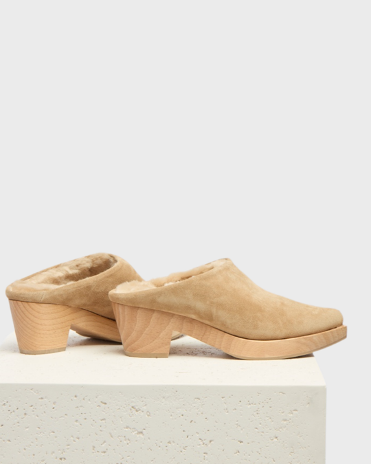 A pair of tan suede clogs with a plush, fur-lined interior, featuring a chunky wooden heel and platform, showcasing a smooth, rounded toe design.