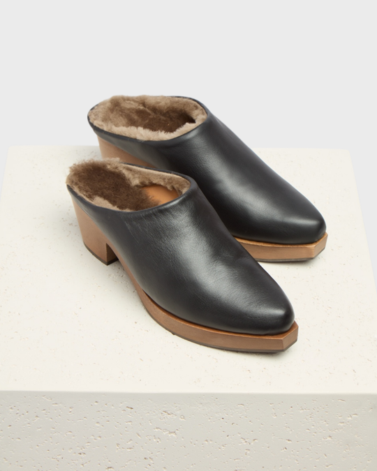 Black leather slip-on clogs with a wooden sole, featuring a plush brown fur lining and a slightly elevated heel.