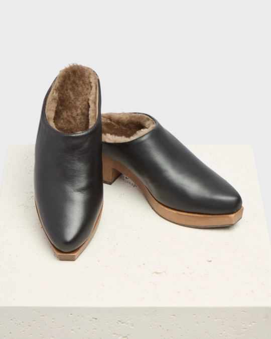 A pair of sleek, black leather slip-on clogs with a smooth finish, featuring a plush brown fur lining, a slightly pointed toe, and a light brown wooden sole with a low heel.