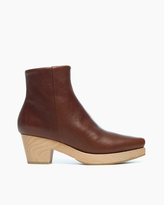 A brown leather ankle boot with a smooth texture, featuring a rounded toe and a light wooden block heel.