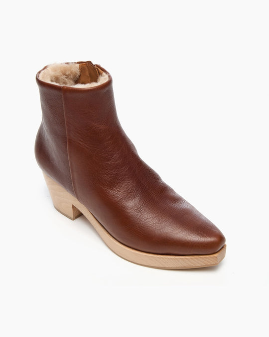 A brown leather ankle boot with a smooth texture, featuring a pointed toe, a wooden wedge heel, and a plush, fur-lined interior.