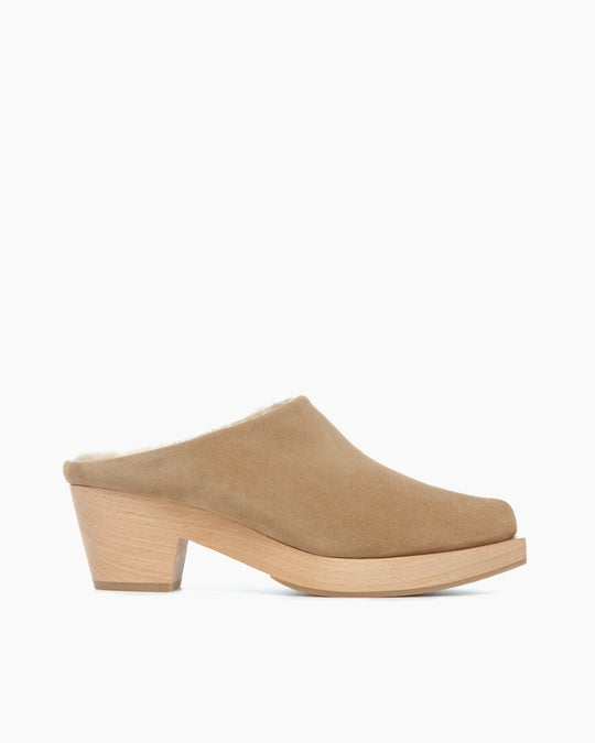 A tan suede mule with a wooden block heel, featuring a rounded toe and a plush, shearling-lined interior.