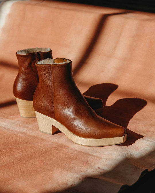 A pair of brown leather ankle boots with a smooth texture, featuring a side zipper, a warm shearling lining, and light wooden block heels.