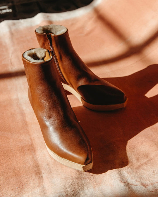 A pair of brown leather ankle boots with a smooth texture, featuring a side zipper, a snipped toe character design, a wooden sole, and a soft, light-colored inner lining.