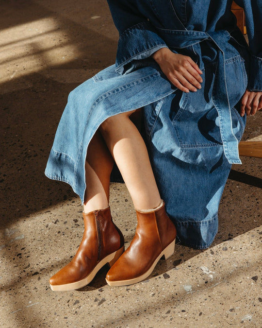 A person wearing a blue denim dress is seated, showcasing a pair of brown leather ankle boots with a smooth finish, tapered toes, and wooden platform heels.