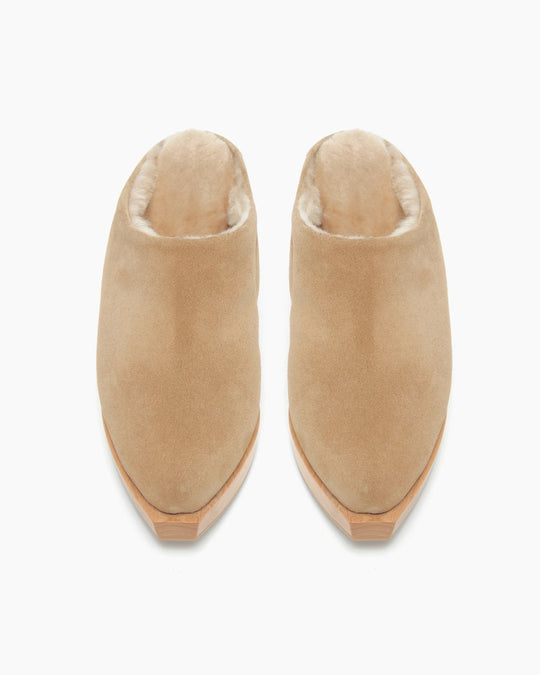 A pair of beige, suede slip-on shoes with a plush, fur-lined interior and wooden soles, featuring a rounded toe and a minimalist design.