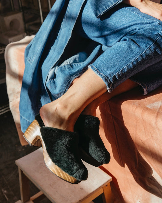 A pair of Hunter Green Shearling slip-on clogs with wooden soles and a slightly elevated heel, worn by a person in blue jeans and a denim jacket, resting on a wooden stool.