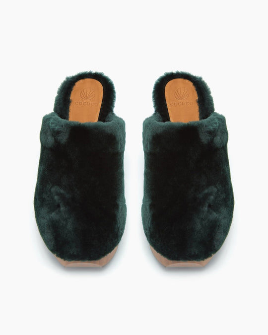 Dark green, plush, slip-on slippers with a soft, fuzzy texture, rounded toe, and a tan sole featuring a logo imprint.