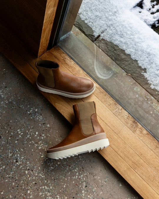 Top view of the France Boot in Caramello in the middle of a wooden doorway with a snowy landscape behind with the left shoe laying flat on the ground.