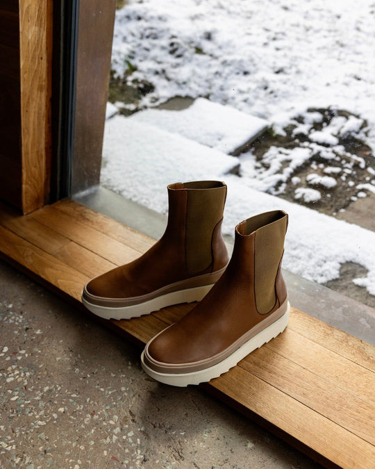 Pair of the France Boot in Caramello in the middle of a wooden doorway with a snowy landscape behind. 