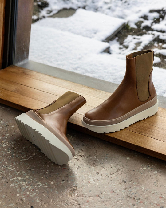 Pair of the France Boot in Caramello in the middle of a wooden doorway with a snowy landscape behind with the right shoe laying flat on the ground.