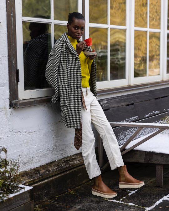 A person wearing a houndstooth coat, a bright yellow sweater, white pants, and tan slip-on shoes with white soles, holding a red cup and standing next to a window with a wooden bench nearby.