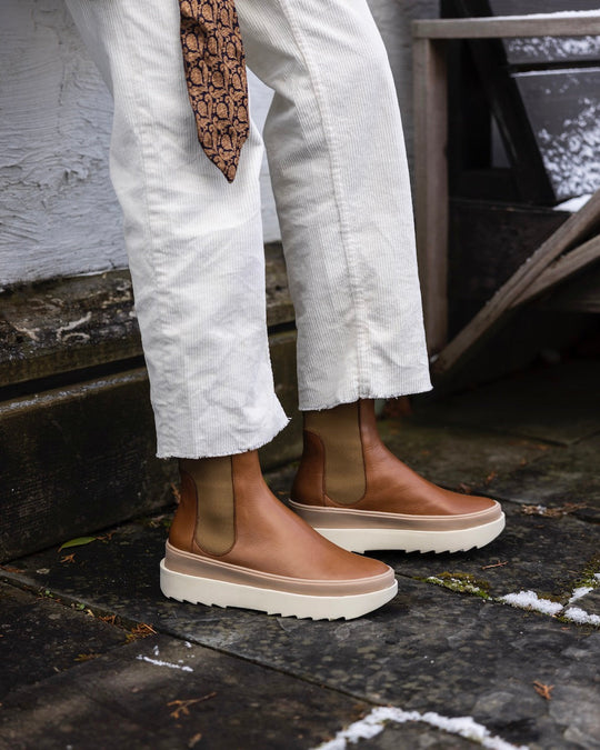 A pair of brown leather Chelsea boots with elastic side panels, white rubber soles, and a sleek, smooth texture, worn with white corduroy pants.