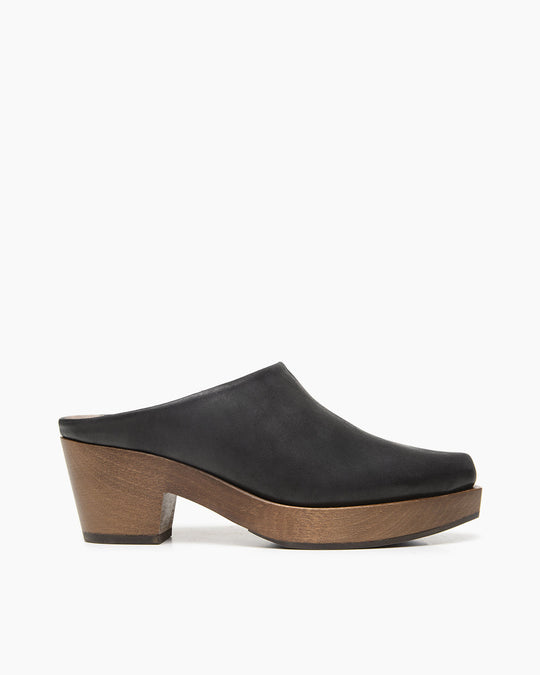 A black leather mule with a smooth texture, featuring a closed toe, a slightly curved upper, and a medium-height wooden platform heel.