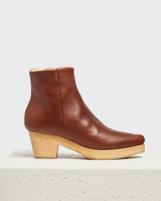 A brown leather ankle clog boot with a smooth texture, featuring a snip toe, a side zipper, a light wooden block heel, and a shearling, cream-colored inner lining.