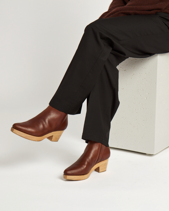 Brown leather clog boots with a smooth texture, featuring a wooden block heel and a slightly snipped toe, worn by a person sitting on a white block.