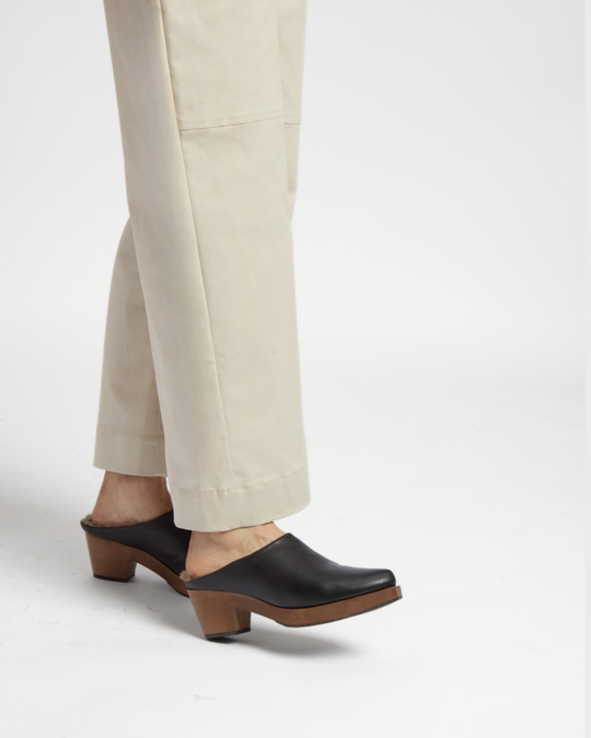 A pair of black leather clogs with a smooth texture, featuring a wooden sole and a chunky heel, worn with beige pants.