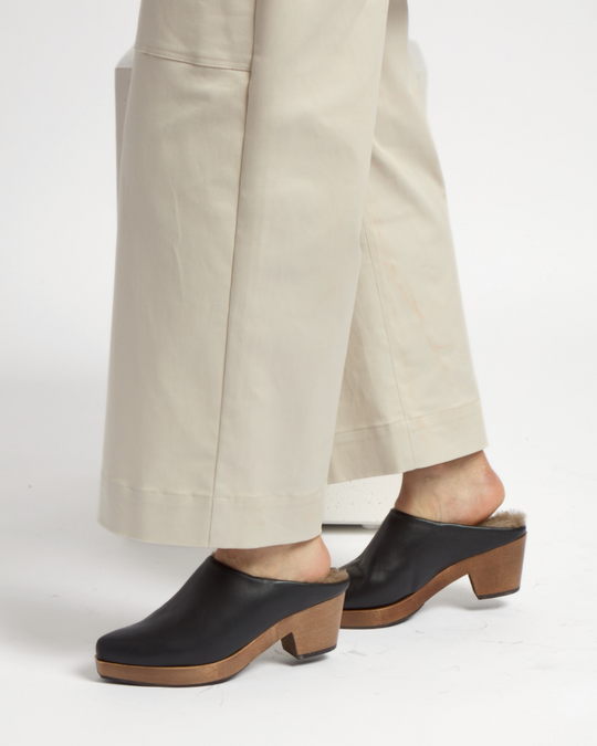 Black leather clogs with a wooden block heel and a soft, fur-lined interior, worn with beige wide-leg pants.