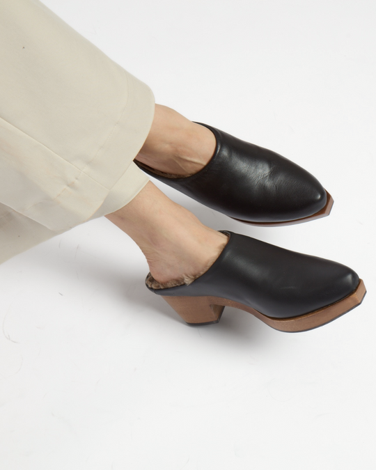 Black leather mule clogs with a smooth texture, featuring a wooden block heel and a shearling-lined interior, worn with light-colored pants.