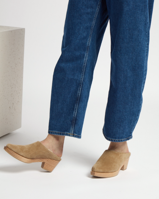 Tan suede clogs with a wooden block heel, featuring a closed toe and a smooth, minimalist design, worn with blue denim jeans.