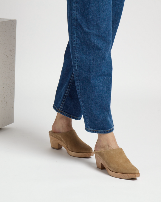 Tan suede mules with a closed toe, wooden block heel, and a smooth, minimalist design, worn with blue denim jeans.