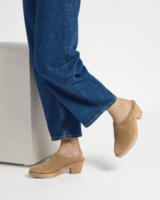 A pair of tan suede clogs with a wooden block heel, featuring a smooth, pointed toe and a slip-on design, worn with blue denim jeans.