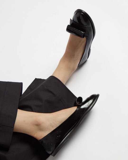 Sleek black patent leather flats with a glossy finish feature a rounded toe and a distinctive bow detail on the front.