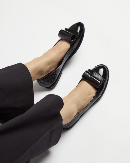 Shiny black leather ballet flats feature a rounded toe, a sleek bow detail on top, and a smooth, glossy finish, complementing their simple, elegant design.