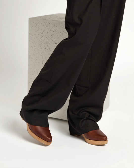 A pair of brown leather clogs with a wooden sole, featuring a sleek, snipped toe design, worn with black, wide-legged pants.